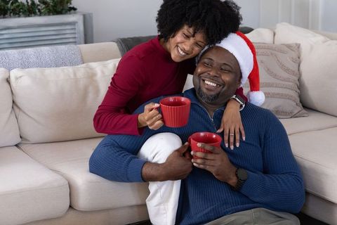 African american couple sharing festive hot drinks on beige sofa wearing santa hat smiling together