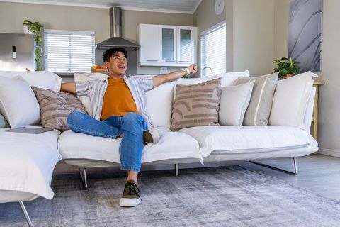 Asian man relaxing on white sectional sofa smiling while using tablet in living room