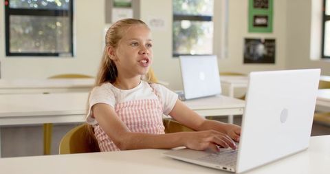 Preteen girl typing and speaking on laptop in bright classroom for online learning