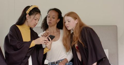 Diverse friends sharing smartphone on bed wearing graduation gowns and casual clothes
