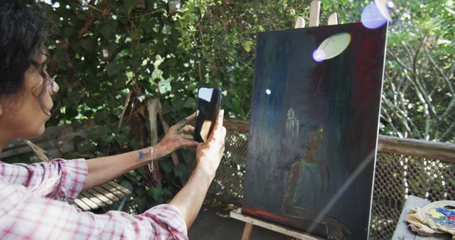 An artist is capturing an image of their dark-themed painting using a smartphone in an outdoor studio. Appealing for use in articles or publications about the artistic process, modern technology in art, self-taught artistry, or promoting art workshops and classes.