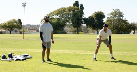 Two teammates practicing cricket on sunny park field with protective gear and stumps
