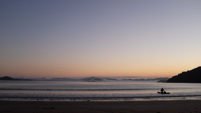 A person is canoeing in the calm ocean near a sandy beach during a beautiful sunset. Mountains are visible in the distance, creating a peaceful atmosphere. Perfect for themes of tranquility, relaxation, adventure, travel, and nature. Ideal for use in travel blogs, adventure magazines, or relaxation ads.