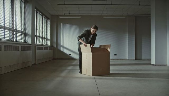 Woman unpacking cardboard box in empty industrial office with blinds casting shadows