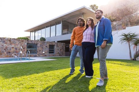 Three friends standing on sunlit lawn at modern hillside home with pool and glass facade