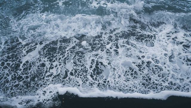 Aerial view of foamy ocean surf forming lacy white patterns over dark pebble shoreline