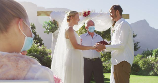 Couple exchanging vows at an outdoor wedding ceremony while wearing face masks. Guests, also wearing masks, witness the ceremony. Useful for topics related to weddings during the pandemic, love in the time of covid-19, safety protocols at events, and social distancing in gatherings.