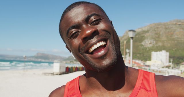 A cheerful man is smiling broadly, enjoying a sunny day on the beach. He wears a red tank top, and behind him are ocean waves, coastal buildings, and colorful beach huts. This image could be used for summer vacation promotions, health and wellness campaigns, positive lifestyle blogs, or advertisements related to travel and leisure.