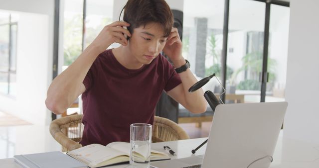 Young Asian man setting up a microphone in home office, wearing headphones, working with a laptop. Great for illustrating remote work, podcasting setup, digital content creation, or tech-savvy lifestyle.