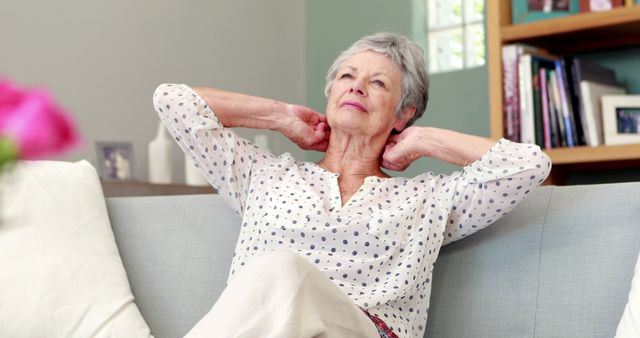 An elderly woman is relaxing on a couch with her hands behind her head. She appears content and comfortable, embodying a casual and serene lifestyle at home. This image is suitable for use in articles or advertisements related to senior living, retirement, comfort at home, and relaxation. It highlights the peace and tranquility of domestic life, making it ideal for blogs, magazines, or campaigns focusing on the well-being and lifestyle of older adults.