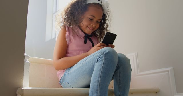 A young girl with curly hair is sitting on the stairs, engrossed in her smartphone. This can be used in articles about children's interaction with technology, family life, or indoor activities.