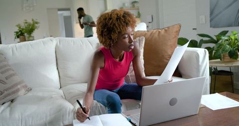 Young woman working from home on couch with laptop and paperwork, man on phone in background