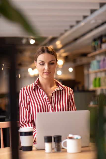 Caucasian businesswoman working on laptop in a coffee shop, wearing a red and white striped shirt. Ideal for illustrating concepts of remote work, business productivity, modern workspaces, and freelance lifestyle. Suitable for use in articles, blogs, and advertisements related to business, technology, and entrepreneurship.