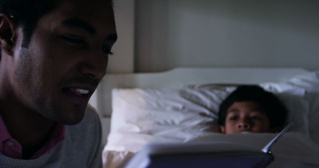 Father reading a storybook to his young son who is lying in bed. Suitable for themes of parenting, family bonding, bedtime routines, and child development. Ideal for use in articles on parenting tips, family life, children's activities, and nighttime routines.