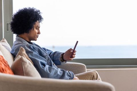 African american man relaxing on sofa by ocean view checking smartphone in denim jacket