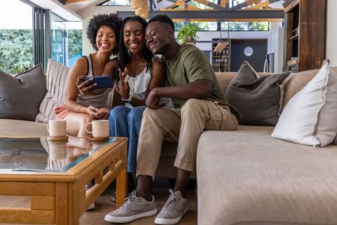 African american friends sharing smartphone and laughing on beige sofa in living room