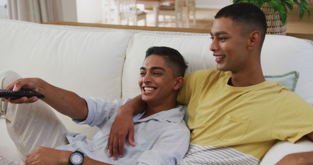 Smiling biracial gay male couple sitting on sofa watching tv one using remote. staying at home in isolation during quarantine lockdown.