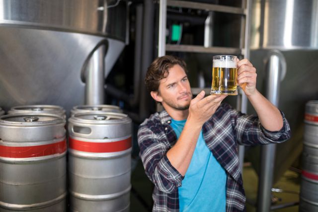 Worker in brewery examining beer quality in glass, surrounded by kegs and brewing equipment. Ideal for use in articles about brewing industry, craft beer production, quality control in beverage manufacturing, and brewery operations.