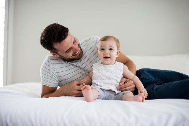 Father and baby girl enjoying quality time together on bed. Perfect for use in parenting blogs, family lifestyle articles, advertisements for baby products, and home decor inspiration.
