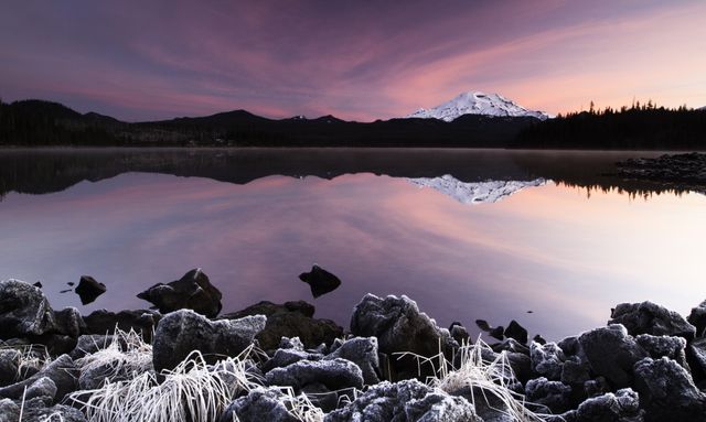 Snow-capped mountain reflecting on tranquil lake at dawn with a pink sky. Foreground has rocky shore and patches of grass, creating a serene and peaceful environment. Ideal for use in travel content, nature promotion, relaxation themes, and scenic desktop wallpapers.