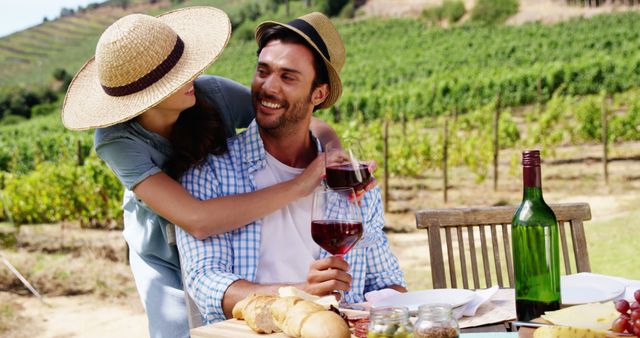 A young Caucasian couple enjoys a romantic picnic with a glass of wine in a vineyard, with copy space. Their cheerful interaction and the scenic backdrop suggest a leisurely day spent in the countryside.