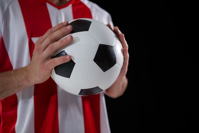Mid-section view of a football player holding a soccer ball with both hands against a black background. The player is wearing a red and white jersey, indicating team colors. This image is ideal for use in sports-related content, advertisements, team promotions, and articles about football or soccer.