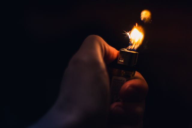 Hand holding lighter creating bright flame in dark environment. Useful for concepts such as lighting, ignition, survival tools, or nightlife. Can be used in industries such as smoking accessories, camping gear, or thriller universe.