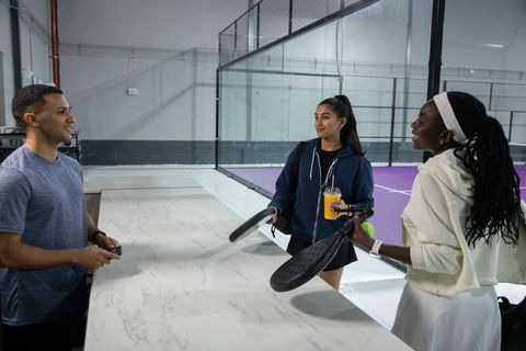 Padel players chatting at marble counter holding rackets and drink beside purple court