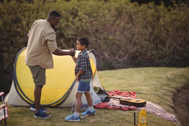 Father and son enjoying a camping trip, shaking hands in front of a yellow tent. Ideal for use in family-oriented advertisements, parenting articles, outdoor activity promotions, and travel brochures.