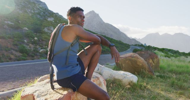 A young man is hiking in the mountains, taking a moment to relax on a large rock by the roadside. He is wearing a tank top, shorts, and a backpack. The scenic mountain view in the background adds to the sense of adventure and outdoor activity. This can be used for promoting active lifestyles, outdoor gear, and tourism.