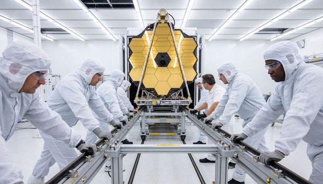 Technicians positioning gold hexagonal space telescope mirror in cleanroom during integration