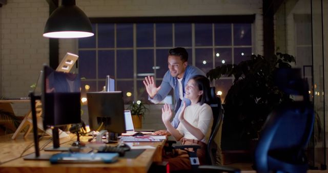 Two colleagues wave at a computer screen while engaging in a video conference during a late-night work session in a sleek, modern office space. Ideal for illustrating topics related to remote work, late-night productivity, business technology, and team collaboration.