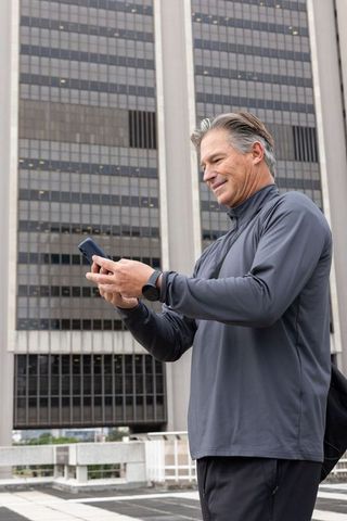 Mature man checking phone on rooftop wearing smartwatch and athletic top downtown highrise