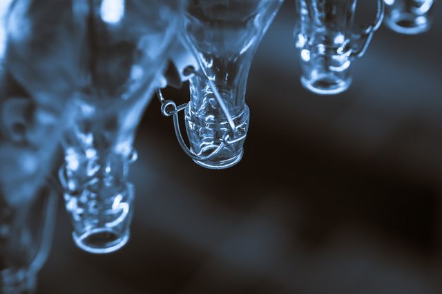 This image captures a close-up view of empty glass bottles hanging upside-down with metal caps, illuminated by soft blue light. The abstract feel evokes a sense of calm and creativity. Useful for backgrounds, abstract designs, and advertising campaigns related to glassware or modern crafts.