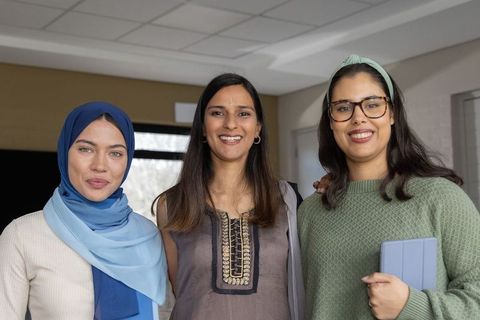 Smiling diverse women collaborating in modern coworking space holding tablet, wearing hijab
