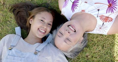 Grandmother and granddaughter are lying on grass, smiling broadly, enjoying each other's company. Both appear relaxed and joyful, embracing a multigenerational connection surrounded by nature. Ideal for illustrating concepts of family unity, generational bonds, and outdoor leisure activities. Can be used in promotions for family events, wellness retreats, or articles on familial relationships.
