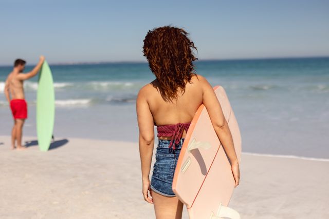 Biracial woman and Caucasian man, both colleagues, are at beach. She has curly brown hair, denim shorts, holding pink surfboard; he's in red shorts, holding a green one, unaltered