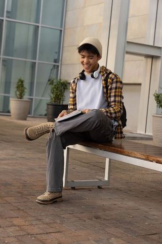 Smiling asian male student sitting on campus bench writing in notebook with headphones