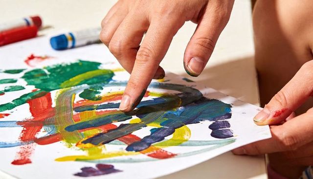 Preschool child finger painting with colorful bright paint strokes and messy hands at table