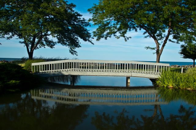 White pedestrian bridge spanning over clear reflection pond surrounded by lush green trees. Ideal for concepts of tranquility, peace, nature, and relaxation. Perfect for travel brochures, nature magazines, blog images about peaceful outdoor spaces, or relaxation-themed content.