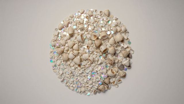 Forming circular pile of pale seashells and holographic sequins on cream tabletop