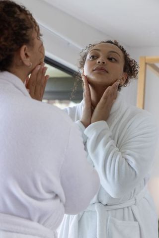 African american woman wearing white robe checking neck and skincare in bathroom mirror