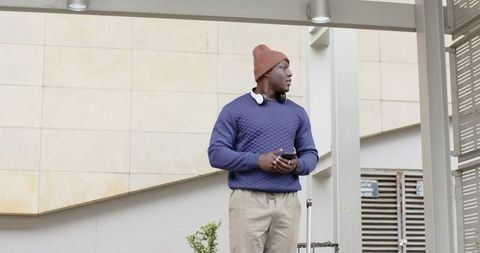 Young african american traveler in rust beanie standing at canopy holding smartphone and luggage