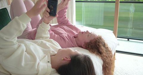 Two friends relaxing on bed in loungewear sharing smartphone near sunlit glass balcony door