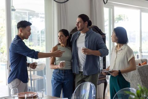 Multicultural friends chatting in bright modern living room holding coffee and tablet