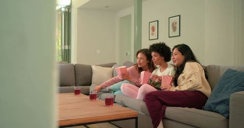 Three friends watching movie night on cozy modern sofa sharing popcorn and drinks
