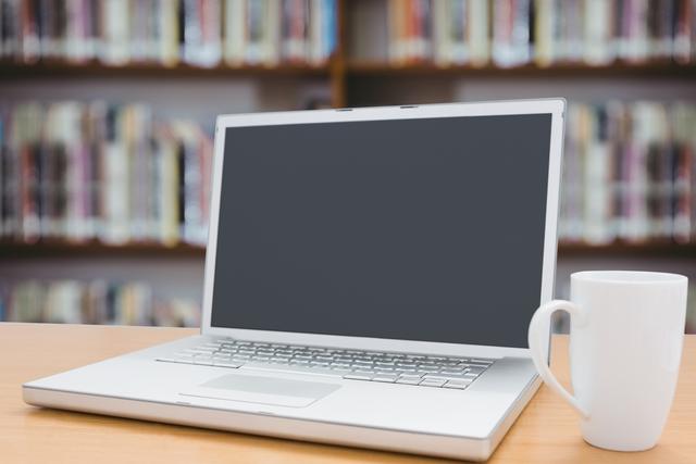 This image shows a laptop and a coffee mug on a desk with a library background. It is ideal for use in educational content, articles about studying or working in libraries, and promotional materials for academic institutions. It can also be used in blogs or websites focused on productivity, technology in education, or student life.