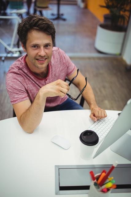 Young male executive smiling while working at his desk in a modern office. He is holding glasses and has a coffee cup nearby. Ideal for use in business, productivity, and workplace-related content.