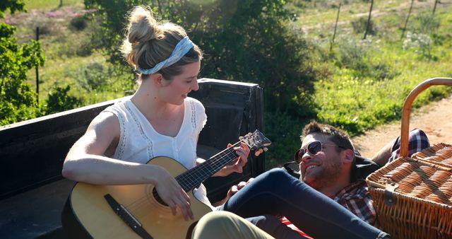 Caucasian couple enjoys a sunny day outdoors, with copy space. She plays the guitar for him in a relaxed, romantic outdoor setting.
