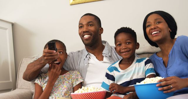 This depicts an African American family enjoying TV time while eating popcorn in their living room. They are all smiling and spending quality time together, suggesting strong family bonds and joyful moments. Suitable for use in family-oriented advertisements, articles on family activities, or any media showcasing togetherness and happiness in a home environment.